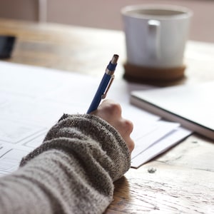 May include: A person's hand holding a blue pen writing on a white sheet of paper on a wooden table. A white coffee mug and a notebook are in the background.