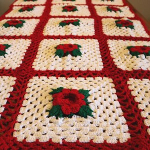 May include: A rectangular crochet table runner featuring a repeating pattern of white granny squares. Each square contains a central three-dimensional red rose motif with green leaves. The squares are joined by a thick, vibrant red yarn border.