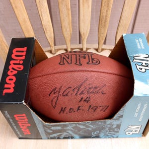 May include: A brown leather Wilson NFL football featuring a black ink autograph and the inscription 14 H.O.F. 1971. The football is housed in its original black and light blue cardboard packaging with the Wilson and NFL logos printed on the sides.