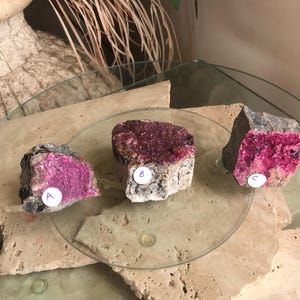 May include: Three raw mineral specimens with a mix of gray and tan rock matrix and vibrant magenta crystal formations. Each specimen is labeled with a letter or number on a white sticker. The minerals are displayed on a glass surface.