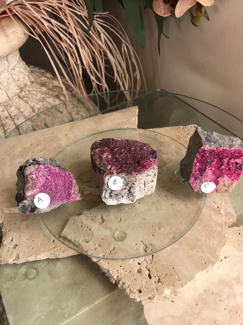 May include: Three raw mineral specimens with a mix of gray and tan rock matrix and vibrant magenta crystal formations. Each specimen is labeled with a letter or number on a white sticker. The minerals are displayed on a glass surface.