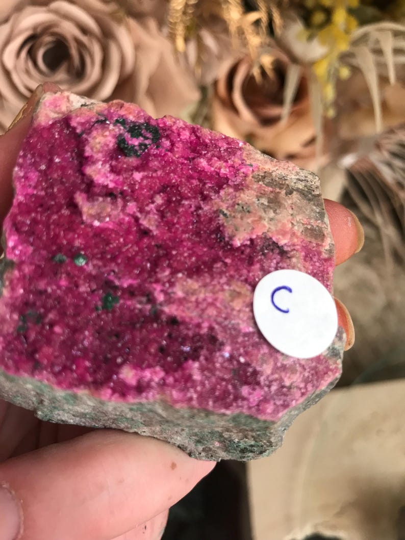 May include: A vibrant, raw mineral specimen featuring a striking combination of colors. The main body displays a rich, hot pink hue, contrasted by areas of green and a neutral, earthy tone. A small white sticker with the letter "C" is visible.