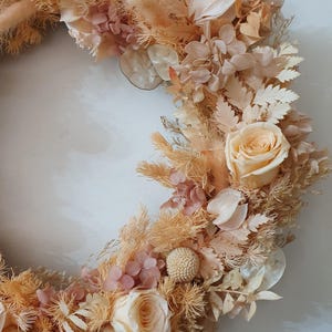 May include: A circular wreath composed of dried and preserved flowers in shades of cream, beige, and soft peach. The arrangement features roses, hydrangea clusters, feathery grasses, and round seed pods, creating a textured floral display.