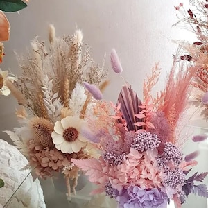 Pode incluir: Dois buquês de flores secas em tons de bege, rosa e roxo. Os arranjos incluem capim-dos-pampas, hortênsias e outros elementos texturizados. Um buquê apresenta uma grande flor branca semelhante a uma margarida.