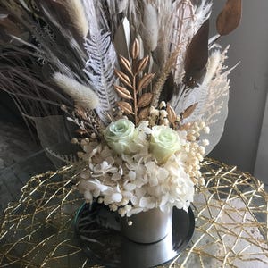 Puede incluir: Arreglo floral con hierba de la pampa seca, helechos y hojas doradas. El arreglo incluye hortensias blancas y dos rosas verde claro en un jarrón plateado. El jarrón está sobre una superficie espejada, sobre una mesa dorada tejida.