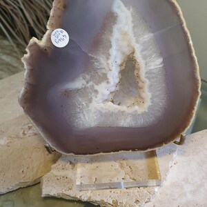 May include: A polished agate geode slice, semi-circular in shape, with bands of lavender, brown, and white. The geode is mounted on a clear acrylic stand, revealing a crystalline centre. The label indicates dimensions of 18 x 15 cm.