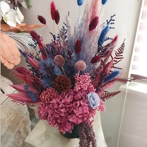 Puede incluir: Un vibrante arreglo floral con una mezcla de flores secas en tonos rosa, burdeos, azul y beige. El ramo incluye hortensias, hierba de la pampa y otros elementos texturizados, creando una composición visualmente rica y en capas. El arreglo está en un jarrón negro.