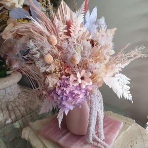 May include: A floral arrangement featuring dried and preserved botanicals in shades of blush pink, lavender, and cream. The bouquet includes pampas grass, billy balls, hydrangea, and a fan-shaped palm leaf, all displayed in a matte pink ceramic vase.