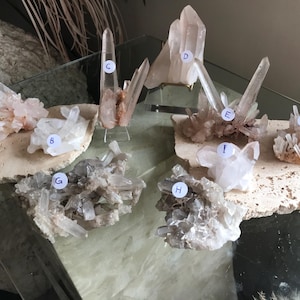 May include: Assortment of quartz crystal clusters in various shapes and sizes. The crystals range in color from clear to pale pink and brown. Some are mounted on a beige stone base, while others stand alone. The crystals are labeled with letters.