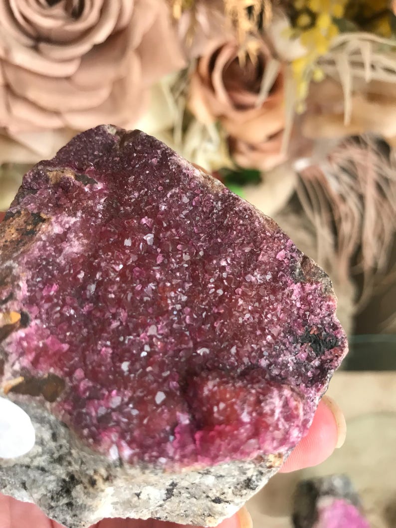 May include: A close-up of a raw mineral specimen featuring a vibrant, crystalline surface in shades of deep magenta and pink. The mineral sits atop a base of gray and white rock, showcasing its natural texture and color variations.