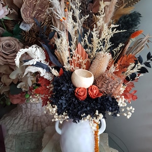 May include: A white ceramic vase with handles overflowing with a dried floral arrangement. The arrangement features a variety of textures and colors, including black, orange, cream, and brown. The bouquet includes roses, grasses, and other dried elements.