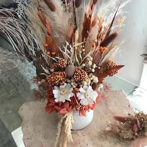 May include: A dried floral arrangement in a white ceramic vase. The bouquet features a variety of textures and colors, including white, orange, brown, and beige. The arrangement includes pampas grass, dried flowers, and other natural elements. A small, matching arrangement sits to the right.
