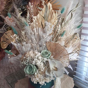 May include: A floral arrangement in a dark teal pot featuring two pale green roses, a white orchid, white hydrangea, dried palm leaves, gold-toned ginkgo leaf accents, and light blue bunny tail grass stems arranged in a tall, textured display.