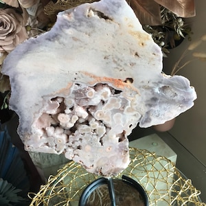 Könnte beinhalten: Eine große, unregelmäßig geformte Geodenscheibe mit helllila und weißem Äußeren. Das Innere zeigt eine Ansammlung kleiner, runder, hellrosa Kristalle. Die Geode ist auf einem schwarzen Metallständer mit goldfarbener Drahtbasis montiert.