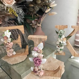 Könnte beinhalten: Drei hölzerne Buchstaben E, I und K, die auf strukturierten Steinsockeln stehen. Jeder Buchstabe ist mit einem Arrangement aus künstlichen Blumen in Weiß-, Creme-, Rosa- und Salbeigrüntönen verziert, mit verschiedenen Blütenformen und Blättern.