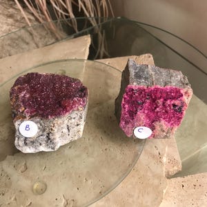 May include: Two rough-cut mineral specimens with vibrant magenta crystal formations. One is labeled "B" and the other "C". The minerals sit on a clear glass surface, with a neutral background.