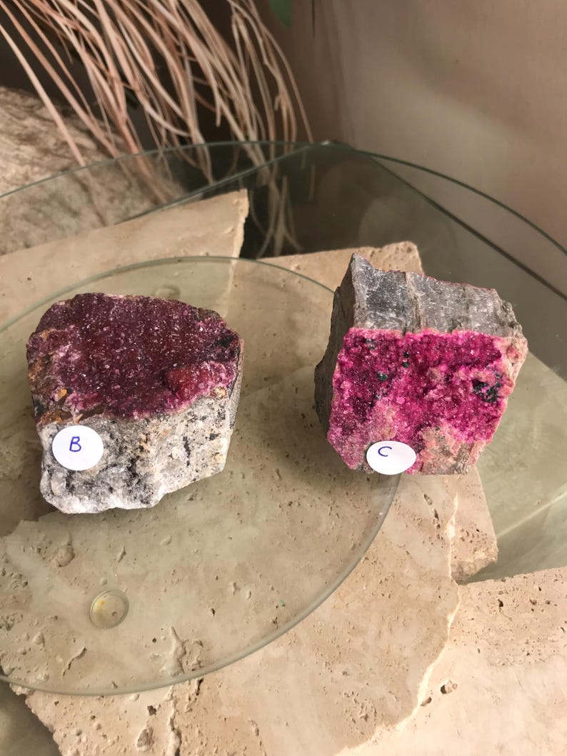 May include: Two rough-cut mineral specimens with vibrant magenta crystal formations. One is labeled "B" and the other "C". The minerals sit on a clear glass surface, with a neutral background.