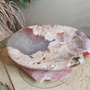 May include: A circular, polished agate slice featuring a marbled pattern of translucent gray, soft pink, and creamy white tones. The stone has a smooth, glossy surface with natural crystalline textures and irregular edges, resting on a pink quartz base.