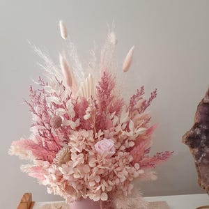 May include: A pink floral arrangement in a round pink vase. The arrangement features dried flowers and grasses in shades of pink, cream, and white. A single pink rose is visible. The vase sits on a light-coloured surface.
