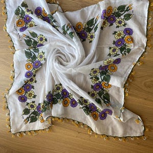 May include: White sheer scarf with a floral pattern in shades of yellow, purple, and green. The scarf has a crocheted border with yellow and white tassels.