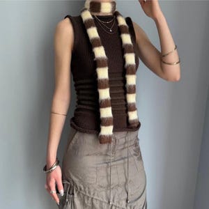 May include: A brown sleeveless top and a matching striped scarf. The scarf features alternating brown and cream-coloured stripes. The outfit is paired with a light brown skirt. The top has a ribbed texture.