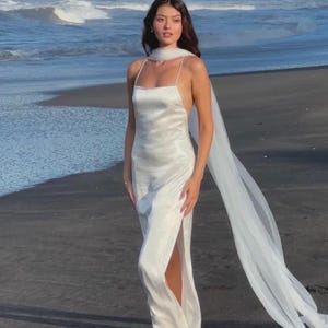 May include: A white satin slip dress with spaghetti straps and a high slit. A sheer white scarf drapes over the shoulders. The dress is worn on a beach with the ocean in the background.