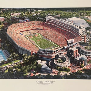 May include: An aerial view of a packed football stadium with a green field and orange seats. The stadium is surrounded by trees and buildings. The text "THE SWAMP" is printed below the image.