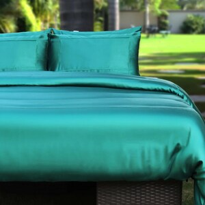 Könnte beinhalten: Ein türkisfarbenes Bettwäsche-Set mit zwei Kissenbezügen und einem Bettbezug. Die Bettwäsche hat eine seidige, glatte Textur und ist ordentlich angeordnet. Das Bild ist im Freien aufgenommen, mit einem verschwommenen grünen Hintergrund aus Bäumen und Gras.