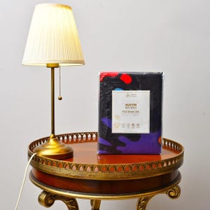 Puede incluir: Una lámpara de mesa con una pantalla plisada y una base dorada se encuentra sobre una mesa redonda y ornamentada. Junto a ella hay un juego de sábanas planas empaquetado con un fondo negro y un diseño abstracto colorido. El embalaje del juego de sábanas tiene el texto "Austin Weird".