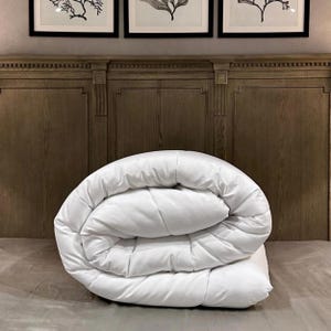 May include: A folded white duvet on a bed. The duvet has a stitched pattern and looks soft and plump. The background includes a wooden headboard and framed pictures.