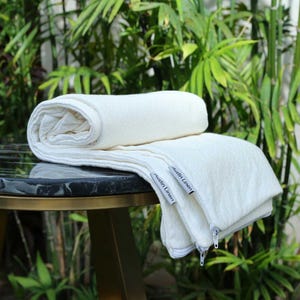 May include: Off-white, rolled and folded blanket on a black marble table. The blanket has a textured surface and a zipper closure. The tag reads "Austin Linen". Green foliage is in the background.