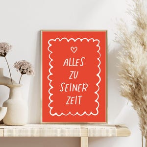 Könnte beinhalten: Ein gerahmter Kunstdruck mit rotem Hintergrund und weißem Text, der "ALLES ZU SEINER ZEIT" in handschriftlicher Form zeigt. Der Text ist von einem weißen, wellenförmigen Rand mit einem kleinen Herzen oben umgeben. Der Rahmen ist goldfarben.