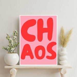 Könnte beinhalten: Gerahmter Kunstdruck mit dem Wort "CHAOS" in großen, roten Buchstaben auf rosa Hintergrund. Der Druck wird auf einem hellen Holzregal präsentiert, mit einer weißen Vase mit Blumen links und einer weißen Vase mit getrocknetem Gras rechts.