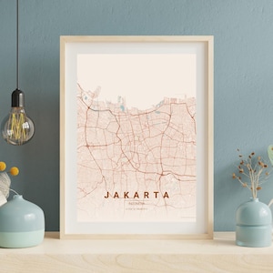 Op de afbeelding: Een ingelijste print van een kaart van Jakarta, Indonesië, in een minimalistische stijl. De kaart toont een netwerk van bruine wegen en infrastructuur tegen een lichtbeige achtergrond. De stadsnaam wordt weergegeven in een vet, bruin lettertype.