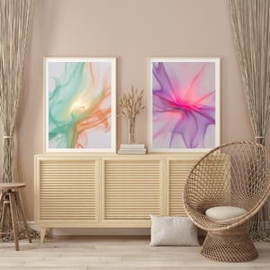 Könnte beinhalten: Zwei gerahmte abstrakte Kunstdrucke. Einer zeigt fließende Türkis-, Orange- und Gelbtöne, während der andere lebendige Rosa- und Lilatöne zeigt. Die Drucke werden über einem hellen Holzschrank mit einem Korbstuhl und neutraler Dekoration ausgestellt.