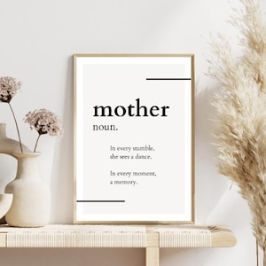May include: A framed print with the word "mother" in a bold, black font, with the definition "noun." and a quote. The print is displayed on a light-colored shelf, with a vase of flowers to the left and decorative dried plants to the right.