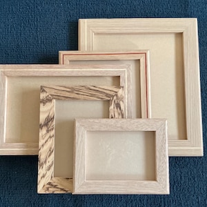 May include: Four wooden picture frames in different sizes. The frames have a natural wood grain finish, with some featuring a contrasting wood pattern. The frames are arranged on a dark blue textured surface.
