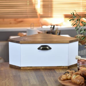 May include: A white wooden bread bin with a triangular, light brown lid and a dark bronze handle. The bin is on a wooden surface, with croissants and honey in the foreground. The bread bin is designed to keep bread fresh.