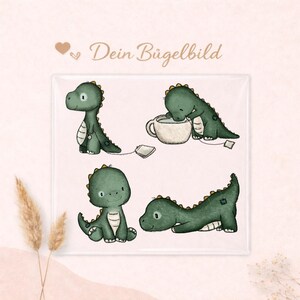 Könnte beinhalten: Quadratisches Bild mit vier Cartoon-Dinosauriern in verschiedenen Posen. Die Dinosaurier sind grün mit gelben Akzenten. Das Bild enthält auch den Text "Dein Bügelbild" in einer dekorativen Schriftart und ein Herzsymbol.