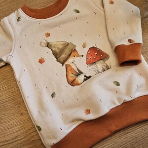 May include: A white long-sleeved sweatshirt with brown trim and a whimsical fox and mushroom graphic. The design includes autumn leaves. The sweatshirt is made of a soft material.