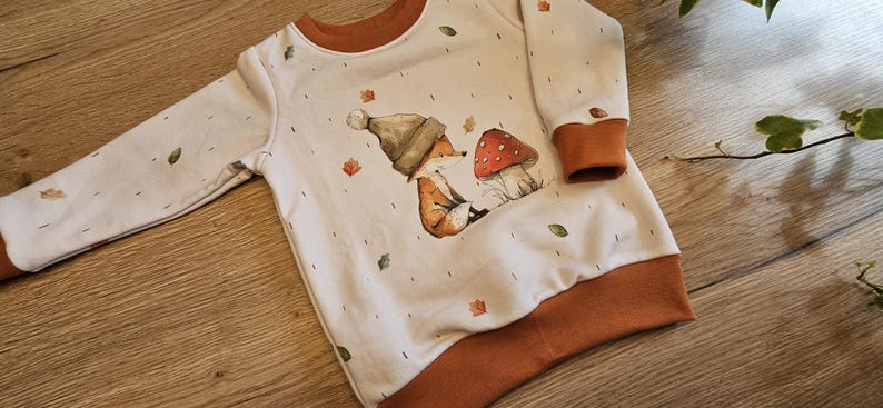 May include: A white long-sleeved sweatshirt with brown trim and a whimsical fox and mushroom graphic. The design includes autumn leaves. The sweatshirt is made of a soft material.