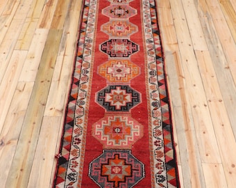 May include: A long, narrow runner rug featuring a vibrant red background with a series of repeating geometric octagonal medallions in shades of orange, pink, grey, and black. The border displays a repeating floral pattern and triangular motifs.