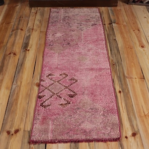 May include: A rectangular, pink runner rug with a faded, distressed appearance. The rug features a geometric design in brown near the bottom. The rug is placed on a wooden floor with visible wood grain.