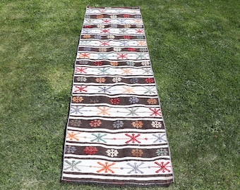 May include: A long, rectangular runner rug featuring alternating horizontal stripes of dark brown and off-white. The white stripes are decorated with a repeating pattern of small, multi-colored star-like floral motifs in red, orange, blue, and gray.
