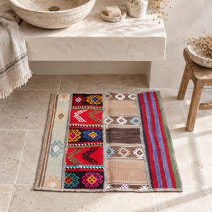 May include: A rectangular woven rug featuring a patchwork design with vertical panels. The rug displays sections of red with geometric patterns in blue, yellow, and green, alongside beige panels with white diamond motifs and a striped section in purple and red.