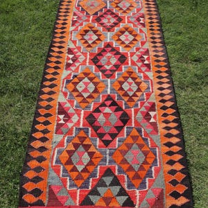 Könnte beinhalten: Ein langer, rechteckiger Teppich mit einem lebendigen geometrischen Muster. Der Teppich hat ein zentrales Design aus Rauten und Dreiecken in Rot-, Orange-, Grau- und Schwarztönen, umrahmt von einem dunklen Rand. Der Teppich liegt auf grünem Gras.