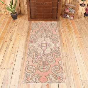 May include: A rectangular runner rug featuring a symmetrical geometric pattern in shades of muted pink, sage green, and cream. The design includes intricate floral-inspired motifs and angular lines arranged in a repeating central medallion layout.