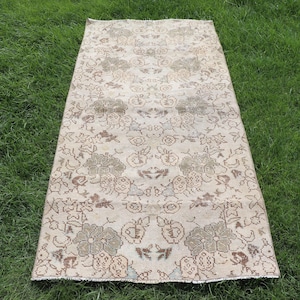 May include: A rectangular, vintage rug with a floral pattern in beige, brown, and green tones. The rug is laid out on a bed of green grass, showcasing its intricate design and aged appearance. The rug's dimensions are not visible.