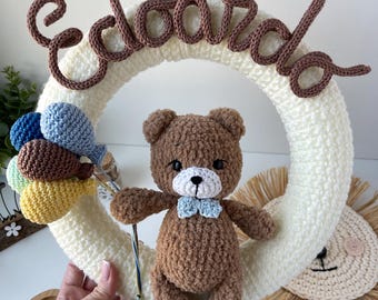 May include: A circular cream-colored crochet wreath featuring the brown yarn-stitched name Edoardo at the top. A brown crochet teddy bear with a light blue bow tie is attached to the center, holding a cluster of small blue, brown, and yellow balloons.