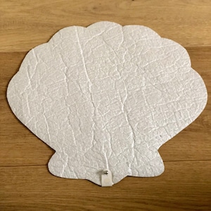 Könnte beinhalten: Ein weißer, muschelförmiger Papierbastelartikel mit einer strukturierten Oberfläche. Die Muschel ist mit einem kleinen weißen Kleber auf einer Holzoberfläche befestigt.